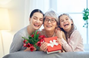 Alt="Happy mother's day! Child and mom congratulating granny giving her flowers tulips ang gift box. Grandma, mum and girl smiling and hugging. Family holiday and togetherness."