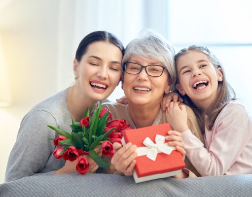 Alt="Happy mother's day! Child and mom congratulating granny giving her flowers tulips ang gift box. Grandma, mum and girl smiling and hugging. Family holiday and togetherness."