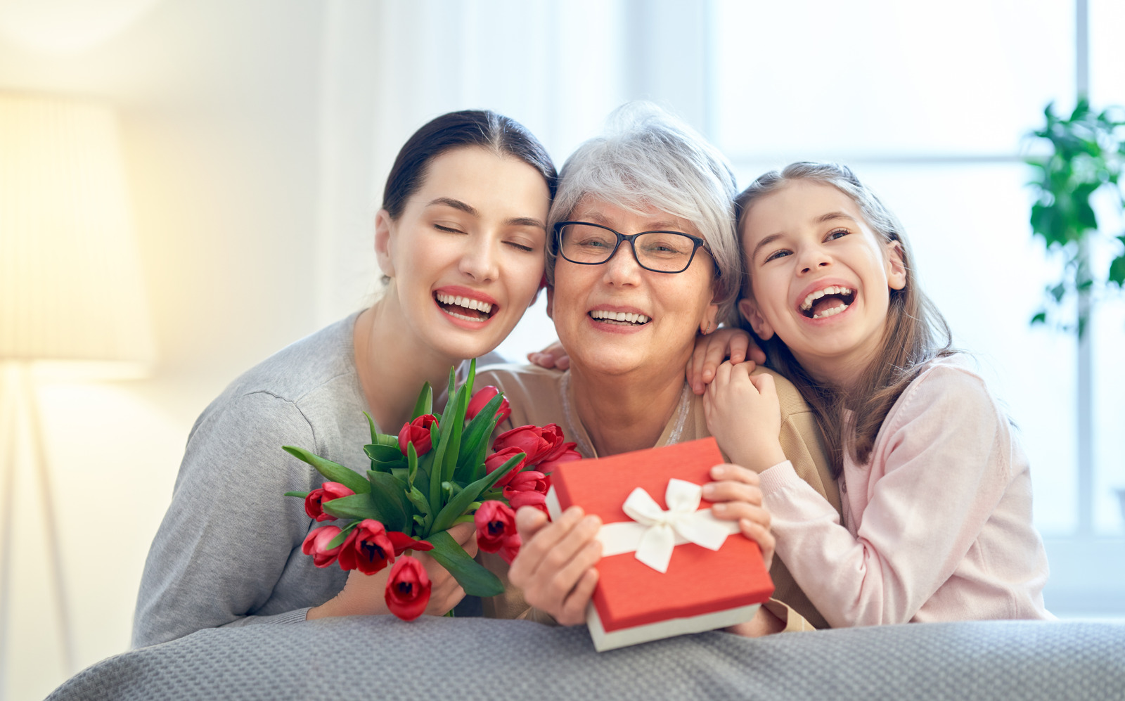 Alt="Happy mother's day! Child and mom congratulating granny giving her flowers tulips ang gift box. Grandma, mum and girl smiling and hugging. Family holiday and togetherness."