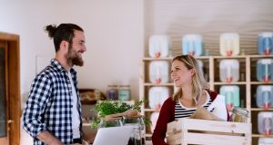 alt=Young male shop assistant with laptop serving an attractive woman in a zero waste shop."