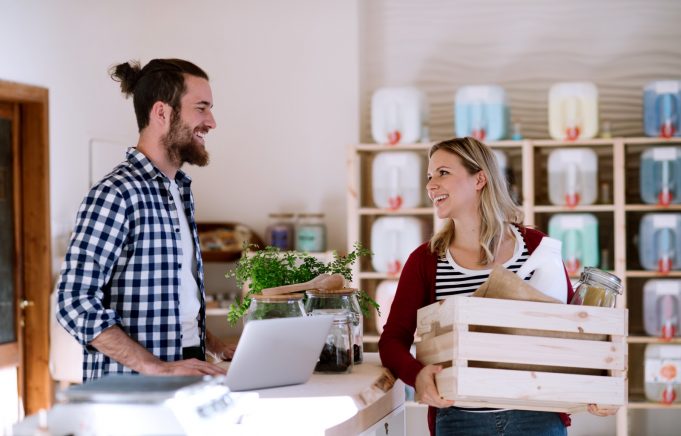 alt=Young male shop assistant with laptop serving an attractive woman in a zero waste shop.