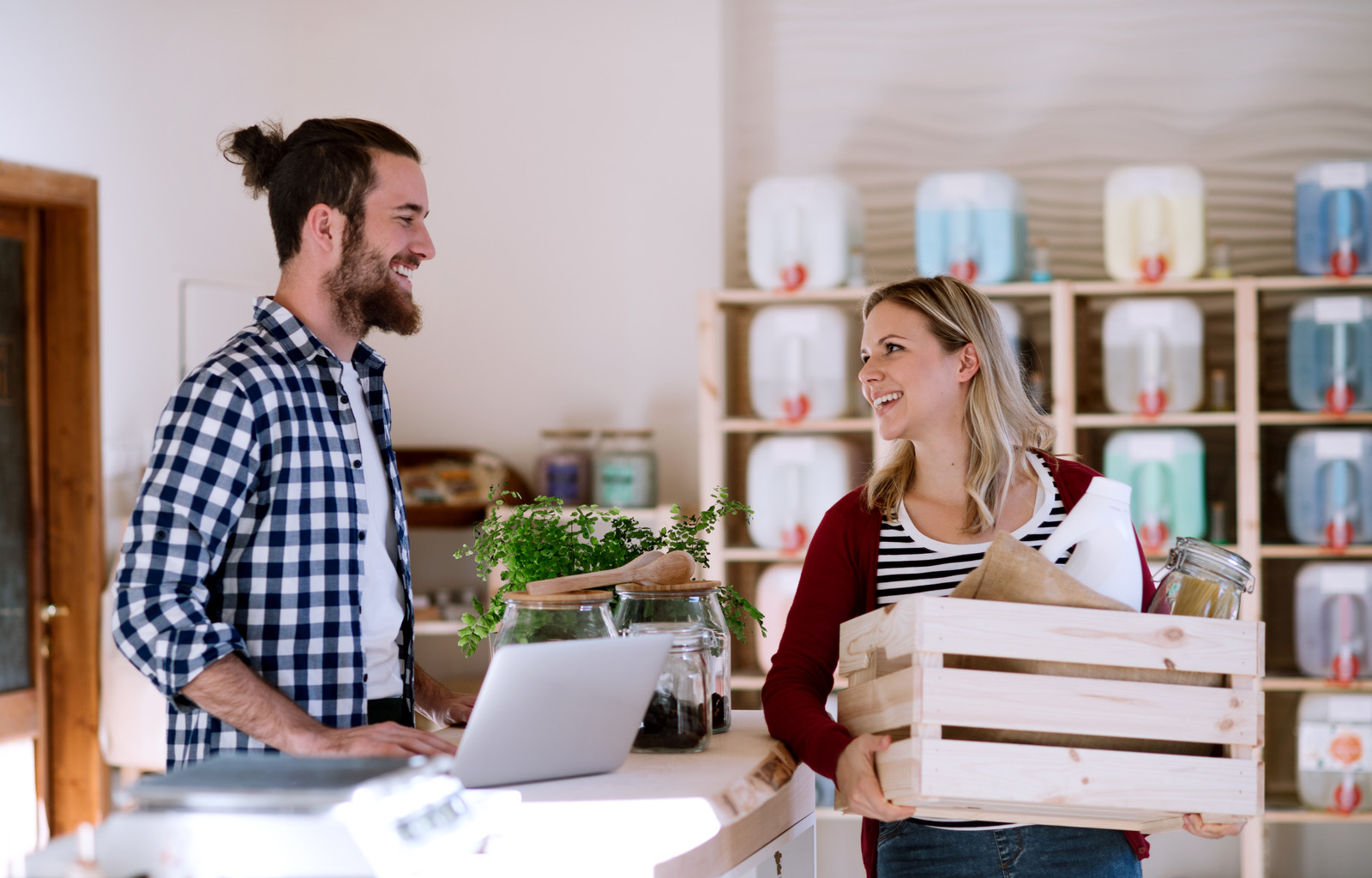 alt=Young male shop assistant with laptop serving an attractive woman in a zero waste shop."