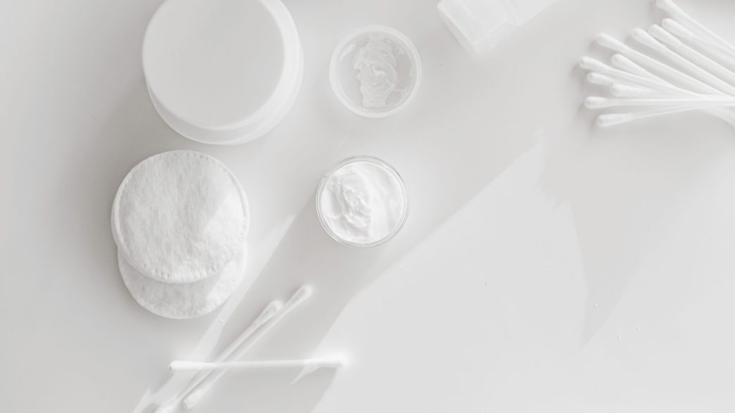 Cotton pads, cotton buds and open jars of face cream on a white surface