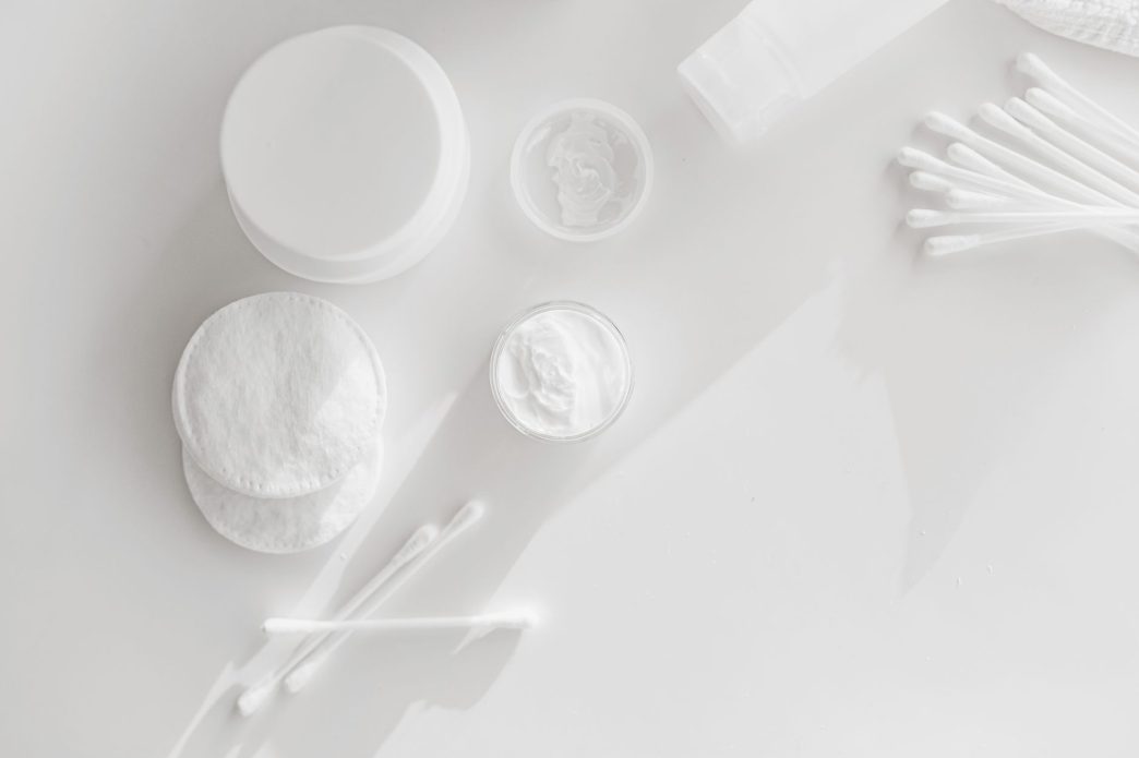Cotton pads, cotton buds and open jars of face cream on a white surface