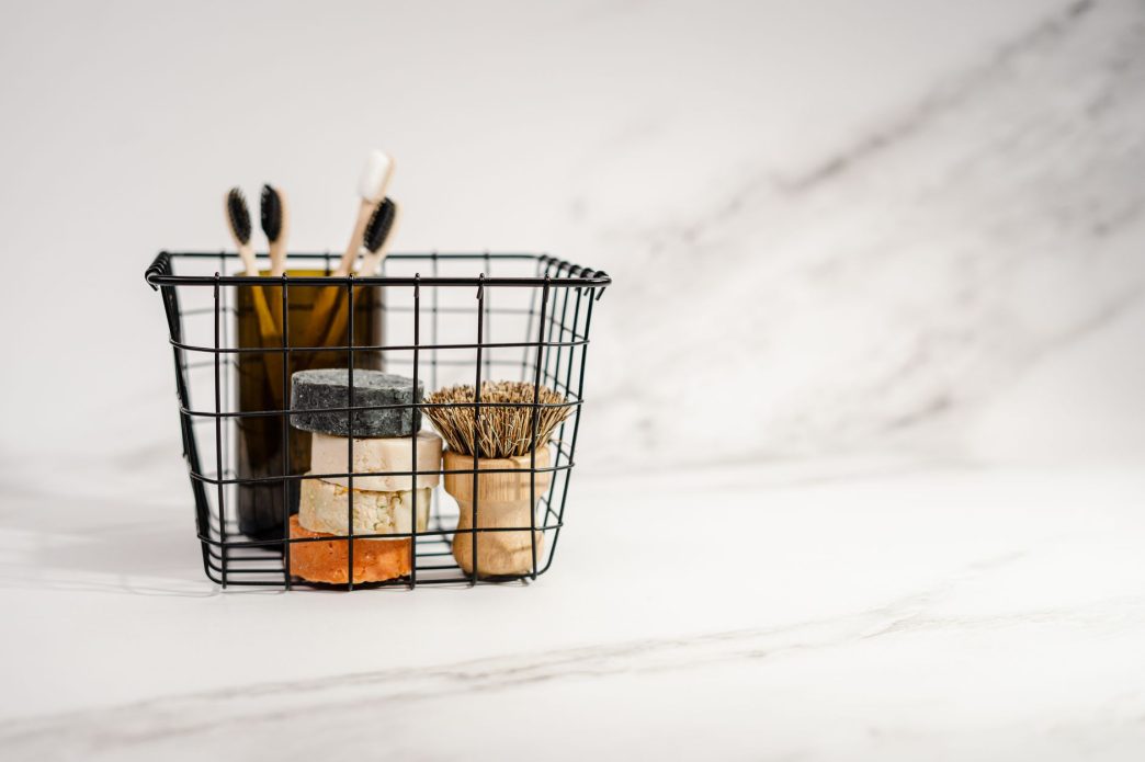 Wire basket holding bars of soap, bamboo toothbrushes and a wooden brush on a white marble surface