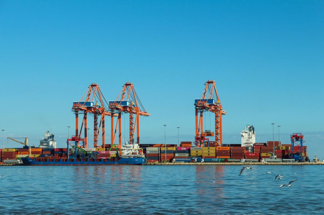 Orange ship-to-shore cranes and stacked shipping containers at a port terminal, with a container ship moored alongside