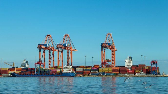 Orange ship-to-shore cranes and stacked shipping containers at a port terminal, with a container ship moored alongside